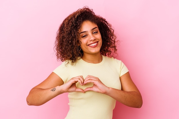 Jovem mulher afro-americana isolada em um fundo rosa, sorrindo e mostrando uma forma de coração com as mãos.