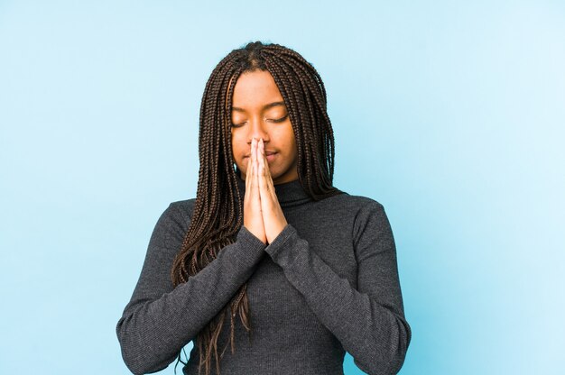 Jovem mulher afro-americana isolada na parede azul, orando de mãos dadas perto da boca, sente-se confiante.
