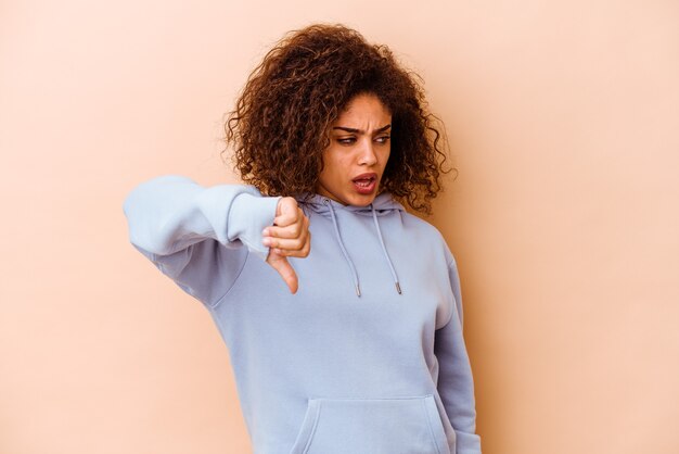 Jovem mulher afro-americana isolada na parede bege, mostrando o polegar para baixo e expressando antipatia.