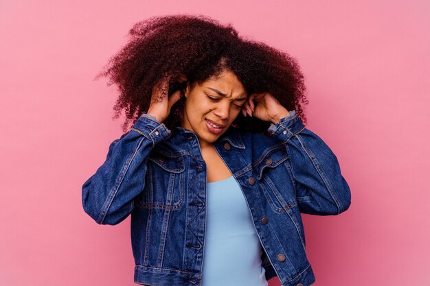 Jovem mulher afro-americana isolada na rosa cobrindo as orelhas com os dedos, estressada e desesperada por um ambiente barulhento.