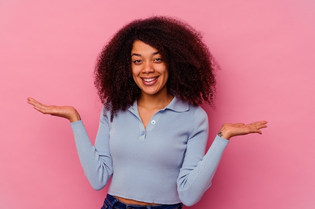 Jovem mulher afro-americana isolada na rosa faz escala com os braços, sente-se feliz e confiante.