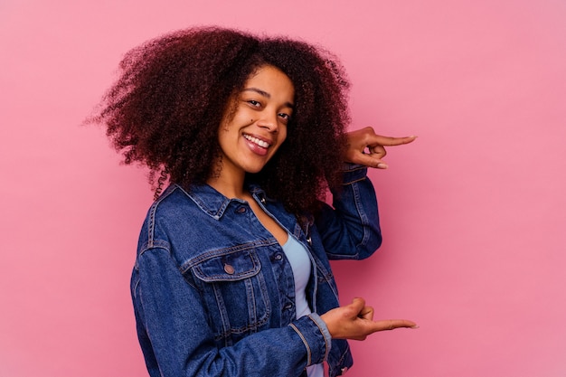 Jovem mulher afro-americana isolada na rosa segurando algo pequeno com os indicadores, sorrindo e confiante.