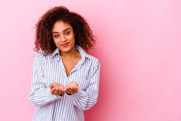 Jovem mulher afro-americana isolada no fundo rosa, dobrando os lábios e segurando as palmas das mãos para enviar beijo no ar.