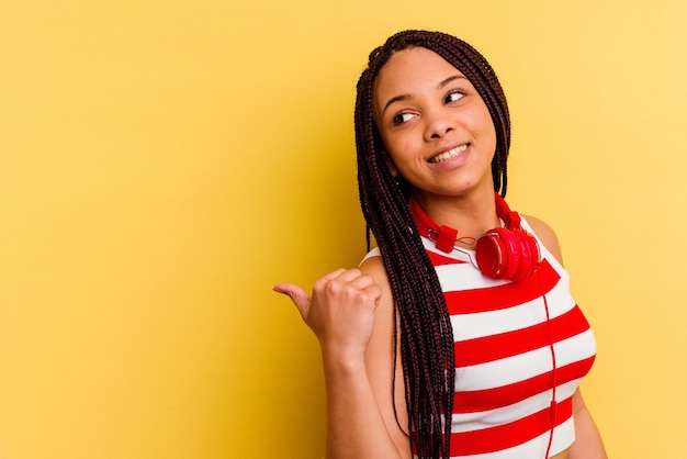 Jovem mulher afro-americana ouvindo música com fones de ouvido isolados na parede amarela aponta com o dedo polegar afastado, rindo e despreocupada.