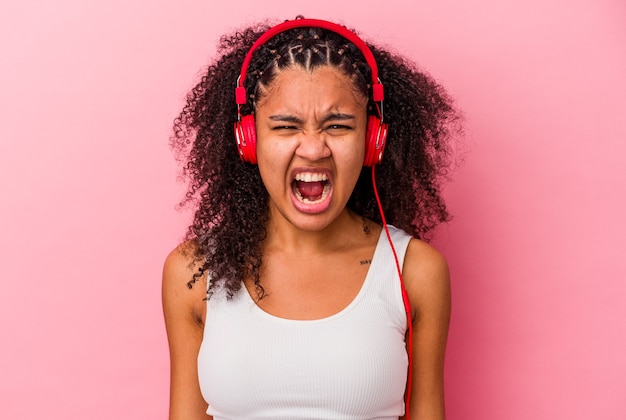 Jovem mulher afro-americana, ouvindo música com fones de ouvido isolados no fundo rosa, gritando muito zangada e agressiva.