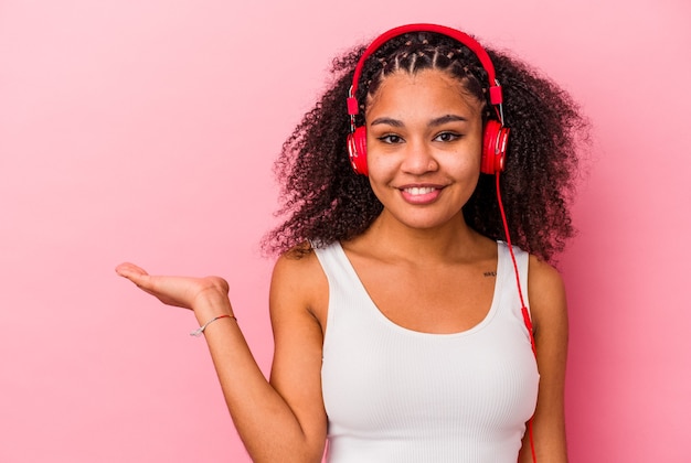 Jovem mulher afro-americana, ouvindo música com fones de ouvido isolados no fundo rosa, mostrando um espaço de cópia na palma da mão e segurando a outra mão na cintura.