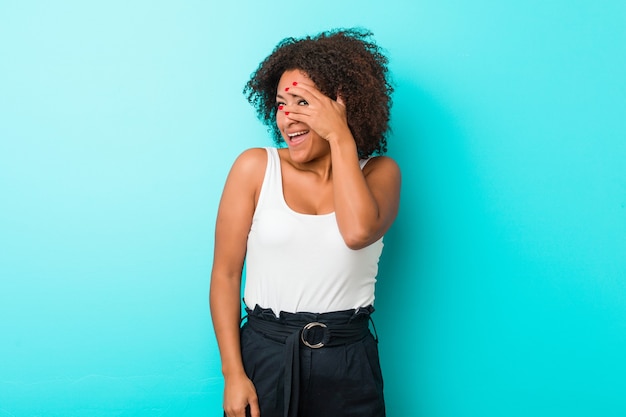 Jovem mulher afro-americana pisca para a câmera por entre os dedos, o rosto coberto de vergonha.