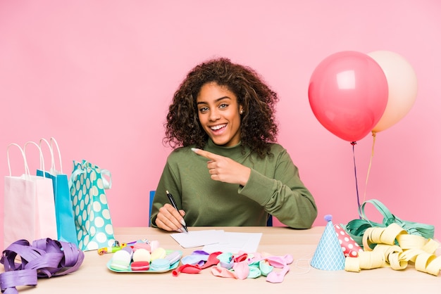 Foto jovem mulher afro-americana planejando um aniversário, sorrindo e apontando de lado, mostrando algo no espaço em branco.