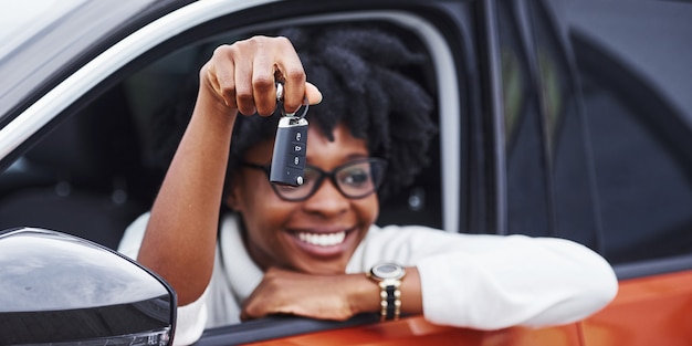 Jovem mulher afro-americana se senta dentro do carro novo e moderno e mostra as chaves.