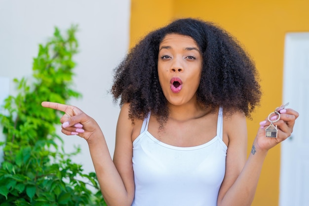 Jovem mulher afro-americana segurando chaves de casa ao ar livre surpreendido e apontando para o lado