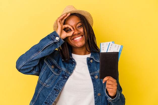 Jovem mulher afro-americana, segurando o passaporte isolado em fundo amarelo, animado, mantendo o gesto ok no olho.