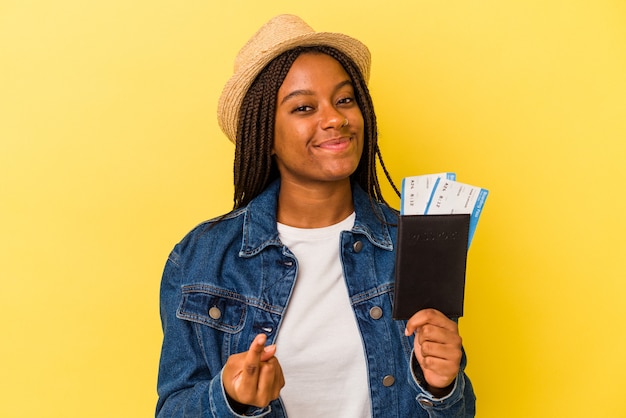 Jovem mulher afro-americana, segurando o passaporte isolado em um fundo amarelo, apontando com o dedo para você como se fosse um convite para se aproximar.