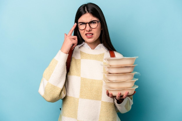 Jovem mulher afro-americana segurando Tupperware isolado em fundo azul, mostrando um gesto de decepção com o dedo indicador