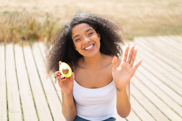 Jovem mulher afro-americana segurando um abacate ao ar livre saudando com a mão com expressão feliz