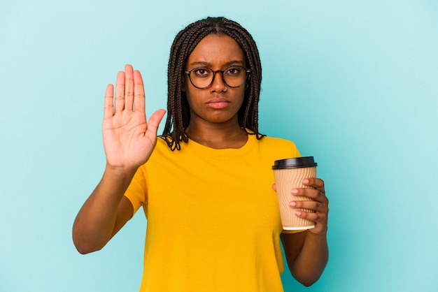 Jovem mulher afro-americana segurando um café para levar isolado em um fundo azul em pé com a mão estendida, mostrando o sinal de pare, impedindo-o.