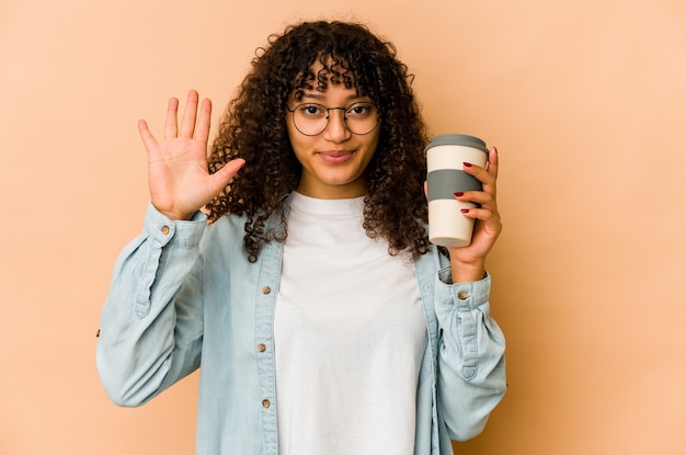 Jovem mulher afro-americana segurando um café para viagem, sorrindo alegre mostrando o número cinco com os dedos.