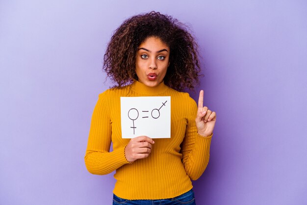 Jovem mulher afro-americana segurando um cartaz de igualdade de gênero isolado na parede roxa, tendo uma ótima ideia, o conceito de criatividade.