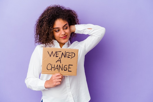 Foto jovem mulher afro-americana segurando um cartaz de mudança, isolado no fundo roxo, tocando a parte de trás da cabeça, pensando e fazendo uma escolha.