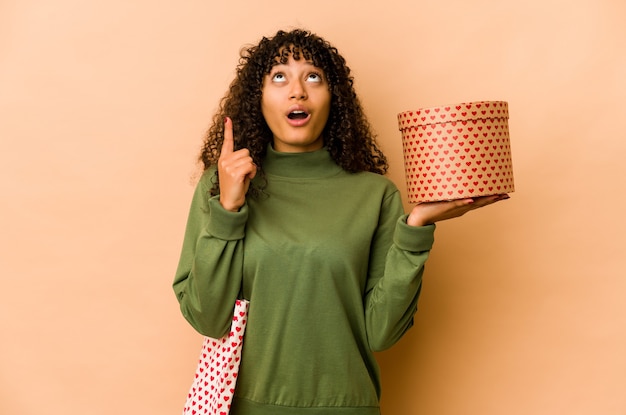 Jovem mulher afro-americana segurando um presente de dia dos namorados apontando para cima com a boca aberta.