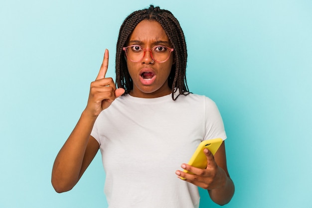 Jovem mulher afro-americana segurando um telefone celular isolado em um fundo azul, tendo uma ideia, o conceito de inspiração.