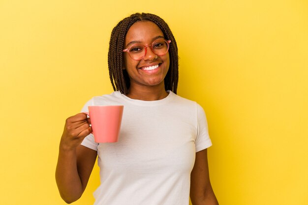 Jovem mulher afro-americana segurando uma caneca isolada em fundo amarelo feliz, sorridente e alegre.