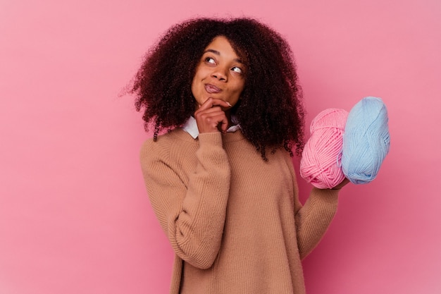 Jovem mulher afro-americana segurando uma linha de costura isolada na rosa, olhando de soslaio com expressão duvidosa e cética.