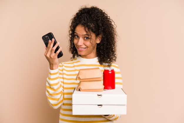 Jovem mulher afro-americana segurando uma pizza e hambúrguer fast-food chamando com telefone celular isolado