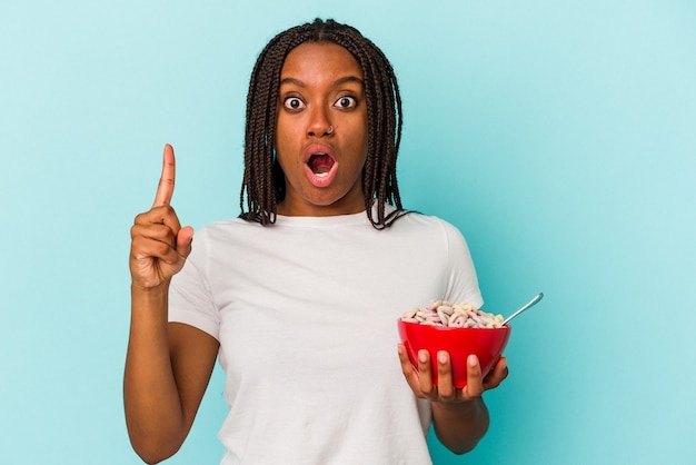 Jovem mulher afro-americana segurando uma tigela de cereales isolada sobre fundo azul, tendo uma ótima ideia, o conceito de criatividade.