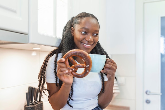 Jovem mulher afro-americana sentada em uma mesa de cozinha e desfrutando de seu pretzel com uma chávena de leite de verme