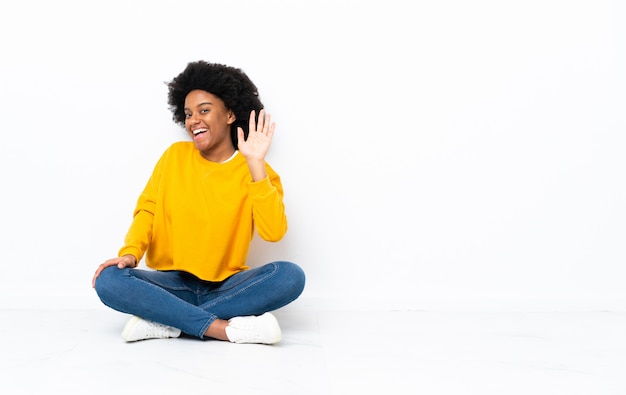 Jovem mulher afro-americana sentada no chão saudando com mão com expressão feliz