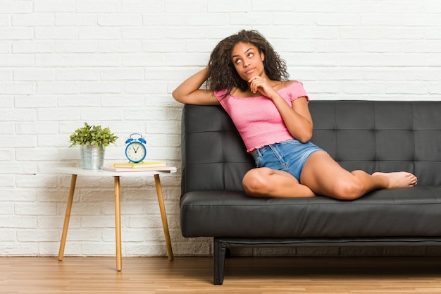 Jovem mulher afro-americana sentada no sofá, olhando de soslaio com expressão duvidosa e cética.