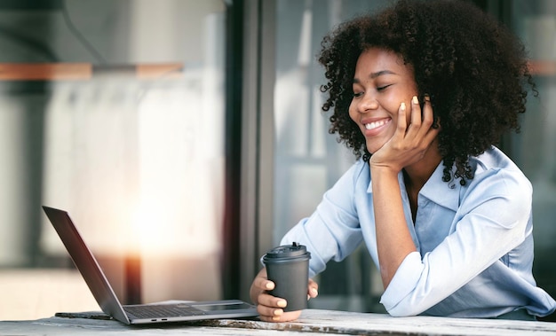 Jovem mulher afro-americana sorridente sentada à mesa no coworkspace olhando para a tela do laptop com felicidade