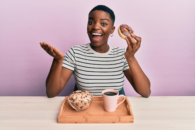 Jovem mulher afro-americana tomando café da manhã segurando rosquinha de chocolate comemorando conquista com sorriso feliz e expressão vencedora com a mão erguida