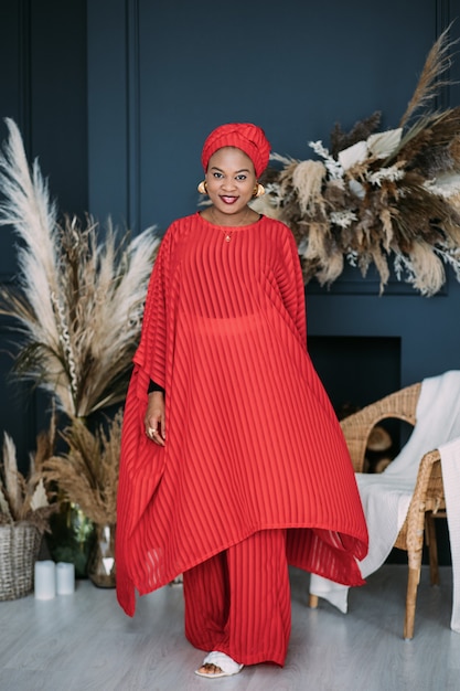 Jovem mulher afro-americana usando um terno brilhante vermelho estilo africano e um xale na cabeça, sorrindo, confiante e feliz, posando em um estúdio moderno