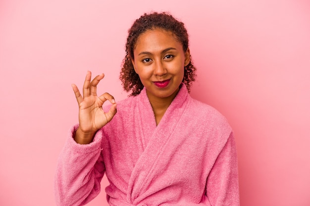 Jovem mulher afro-americana, vestindo um roupão de banho isolado no fundo rosa, alegre e confiante, mostrando um gesto ok.
