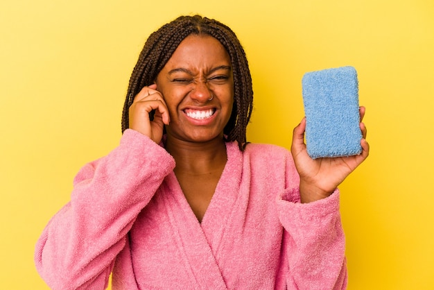 Jovem mulher afro-americana, vestindo um roupão de banho, segurando uma esponja azul isolada em um fundo amarelo, cobrindo as orelhas com as mãos.