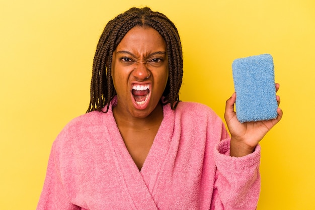 Jovem mulher afro-americana, vestindo um roupão de banho, segurando uma esponja azul isolada no fundo amarelo, gritando com muita raiva e agressividade.
