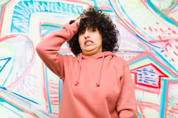 Foto jovem mulher afro bonita entrando em pânico por um prazo esquecido, sentindo-se estressada, tendo que encobrir uma bagunça ou erro contra a parede do graffiti