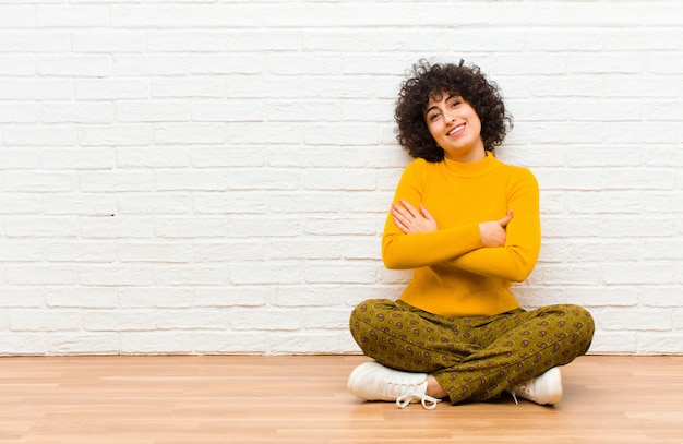 Jovem mulher afro bonita rindo alegremente com os braços cruzados, com uma pose relaxada, positiva e satisfeita, sentada no chão