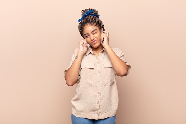 Jovem mulher afro com cara de zangada, estressada e irritada, cobrindo os ouvidos com um barulho, som ou música alta ensurdecedores