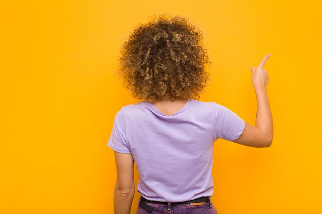 Jovem mulher afro em pé e apontando para objetar no espaço da cópia, vista traseira contra a parede laranja