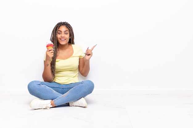 Jovem mulher afro parecendo animada e surpresa, apontando para o lado e para cima para copiar o espaço. conceito de sorvete