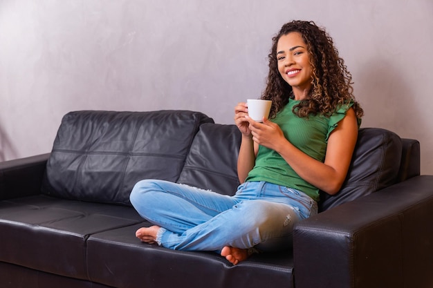 Jovem mulher afro relaxando no sofá, tomando chá ou café em uma xícara.