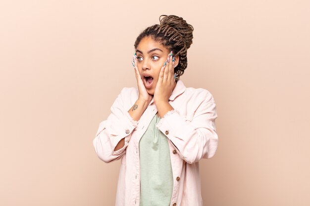 Jovem mulher afro se sentindo feliz, animada e surpresa, olhando para o lado com as duas mãos no rosto