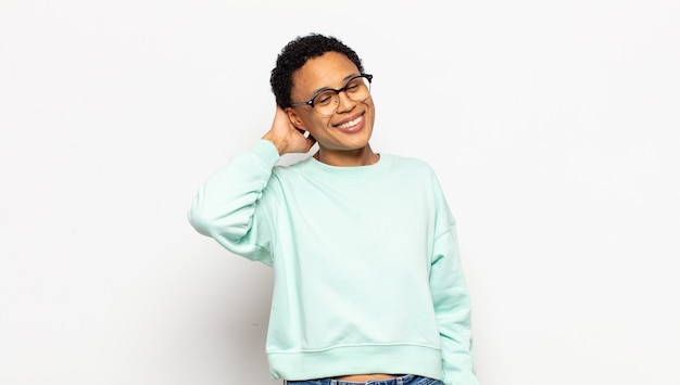 Jovem mulher afro sorrindo alegre e casualmente, levando a mão na cabeça com um olhar positivo, feliz e confiante