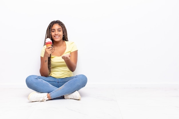 Jovem mulher afro sorrindo alegremente com olhar amigável, confiante e positivo, oferecendo e mostrando um objeto ou conceito. conceito de sorvete