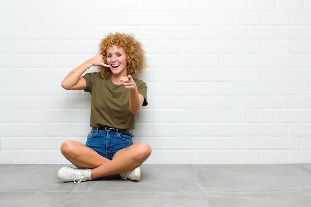 Jovem mulher afro, sorrindo alegremente e apontando para a câmera enquanto faz uma ligação, gesto mais tarde, falando no telefone, sentado no chão