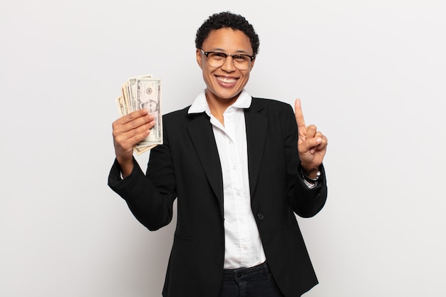 Jovem mulher afro sorrindo com orgulho e confiança fazendo a pose número um triunfantemente, sentindo-se uma líder