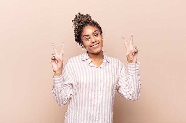 Jovem mulher afro sorrindo e parecendo feliz, amigável e satisfeita, gesticulando vitória ou paz com as duas mãos