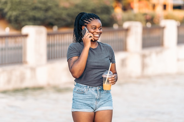 Jovem mulher afro sorrindo enquanto falava com um telefone celular e segura um shake na rua.